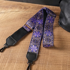 Könnte beinhalten: Ein Ersatz-Taschenriemen mit einem gewebten violett-schwarzen Blumenmuster und metallisch silbernen Akzenten. Der Riemen verfügt über schwarze Lederenden, schwarze drehbare Karabinerhaken aus Metall und eine verstellbare Kunststoffschnalle.