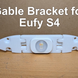 May include: A white plastic gable bracket for Eufy S4, featuring a central light component. The bracket has hinges and mounting holes. The text "Gable Bracket for Eufy S4" is displayed in white.
