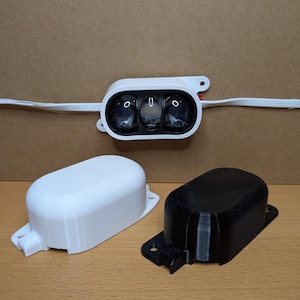 May include: A white plastic device with three clear lenses and a white wire. Below are two rectangular cases, one white and one black, with mounting tabs. The items are on a wooden surface.