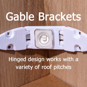 May include: White plastic gable brackets for LED strip lights. The brackets have a hinged design that allows them to be adjusted to fit different roof pitches. The text "Gable Brackets" is printed above the brackets. The text "Hinged design works with a variety of roof pitches" is printed below the brackets.
