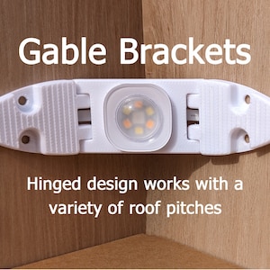 May include: White plastic gable brackets with a hinged design for mounting LED strip lights. The brackets are designed to work with a variety of roof pitches. "Gable Brackets" is written in black text above the brackets. "Hinged design works with a variety of roof pitches" is written in black text below the brackets.