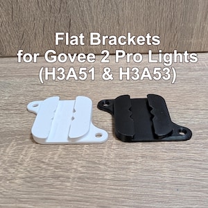 May include: One white and one black flat bracket for Govee 2 Pro Lights (H3A51 & H3A53). Each bracket features a central channel with serrated edges and two circular mounting holes on opposite sides, designed for securing light strips to flat surfaces.