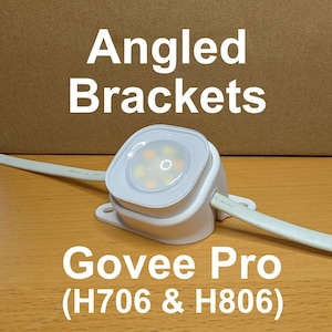 May include: White angled brackets with a square LED light fixture. The text "Angled Brackets" and "Govee Pro (H706 & H806)" are displayed in white. The fixture is on a wooden surface.