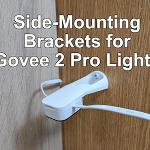 Peut inclure: Un support de montage latéral en plastique blanc pour les lumières Govee 2 Pro, doté d'une base à visser et d'une fente pour la bande lumineuse. Le texte "Side-Mounting Brackets for Govee 2 Pro Lights" est superposé au-dessus du matériel.