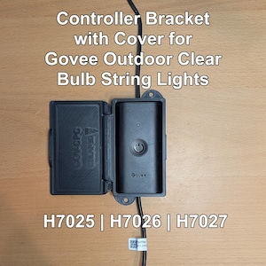 May include: Black controller bracket with cover for Govee Outdoor Clear Bulb String Lights. The bracket is open, revealing a black controller with a power button. Text on the bracket reads "Controller Bracket with Cover for Govee Outdoor Clear Bulb String Lights".