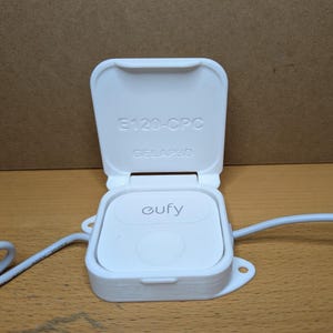 May include: A white Eufy device with a hinged lid, open to reveal the Eufy logo. The lid has the text "E120-CPC" and "GELAP3D". A white cable extends from the device. The device is on a wooden surface.