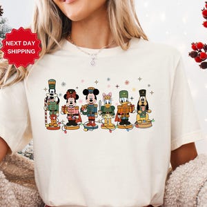 May include: Cream-colored t-shirt featuring a festive design of cartoon nutcrackers. The nutcrackers are dressed in various outfits and are arranged in a row. The shirt also has the text "NEXT DAY SHIPPING".