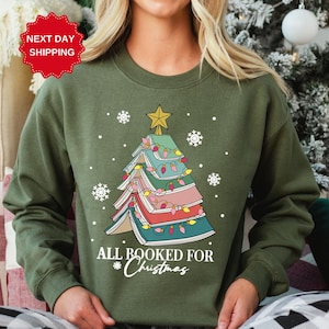 Puede incluir: Sudadera verde con un &aacute;rbol de Navidad hecho de libros apilados con luces. El texto "All Booked for Christmas" est&aacute; impreso debajo del &aacute;rbol.