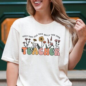May include: A white t-shirt with a floral design and the text "Teach them. Love them. Watch them grow. Teacher" in colorful letters.