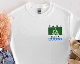May include: White t-shirt with a green and blue camp logo. The logo says "CAMP NAME" with a campfire graphic and wavy lines.