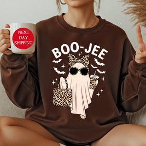May include: Brown sweatshirt with a Halloween-themed graphic. The graphic features a ghost wearing sunglasses, a bow, and holding a leopard print bag and coffee cup. The text "BOO-JEE" is above the ghost. A white mug with red text is held in the hand.