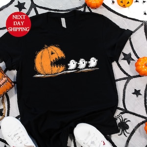 May include: Black t-shirt featuring a Halloween-themed graphic. The design shows an orange pumpkin with a carved face chasing three white ghosts. The shirt is displayed with white sneakers and a decorative purse.