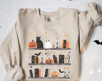 May include: Beige sweatshirt featuring a Halloween-themed bookshelf design. The graphic includes pumpkins, a black cat, a ghost, candles, books, and bats. The design is printed on a soft, comfortable sweatshirt, perfect for the fall season.