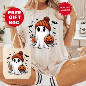Może przedstawiać: Biały t-shirt z uroczym kresk&oacute;wkowym duchem w pomarańczowej czapce, trzymającym dynię i kubek kawy. Duch otoczony jest nietoperzami i gwiazdami. Do koszulki dołączona jest bezpłatna torba.
