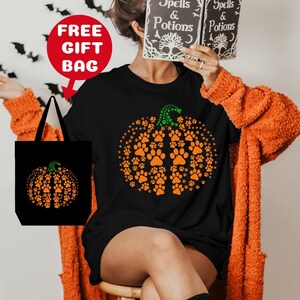 May include: Black t-shirt with a pumpkin design made of orange paw prints. The shirt is paired with an orange cardigan and a black tote bag with the same pumpkin design. The tote bag has the text "FREE GIFT BAG" in red.