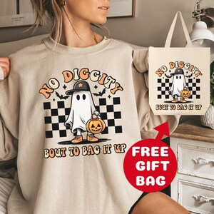 May include: A beige sweatshirt with a graphic of a ghost wearing a hat and holding a pumpkin. The text "NO DIGGITY BOUT TO BAG IT UP" is printed on the sweatshirt. A free tote bag with the same graphic and text is included.