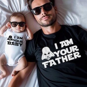 May include: A black t-shirt with the text "I AM YOUR FATHER" and a white Darth Vader graphic, next to a baby in a white onesie with the text "I AM YOUR BABY" and a Darth Vader graphic. Both are wearing black sunglasses.