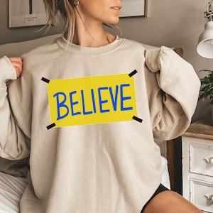 May include: A beige sweatshirt with a yellow rectangle with black tape on the corners. The rectangle has the word "BELIEVE" written in blue letters.