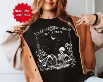 Puede incluir: Camiseta negra con un gráfico blanco de un esqueleto leyendo un libro en un jardín. El gráfico incluye el texto "Read in Peace".
