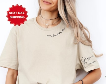 Op de afbeelding: Een beige T-shirt met het woord "mama" in zwarte cursieve letters op de voorkant en de naam "Grayson Conner" in zwarte cursieve letters op de rechtermouw.