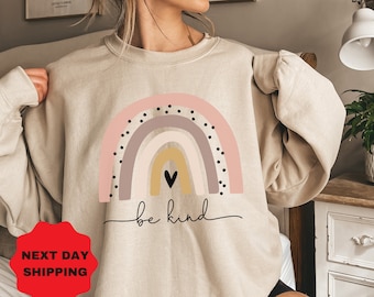 Puede incluir: Una sudadera beige con un gráfico de arcoíris y el texto "be kind". El arcoíris está formado por rayas rosas, moradas, beige y amarillas con lunares negros.