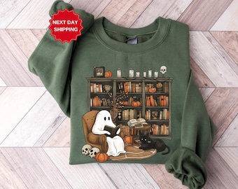 Könnte beinhalten: Ein grünes Sweatshirt mit einem Aufdruck eines Geistes, der in einem Sessel vor einem Bücherregal voller Bücher ein Buch liest. Auf dem Regal befinden sich Kürbisse, Kerzen und Schädel. Der Text "Next Day Shipping" befindet sich auf einem roten Stern-Aufkleber.