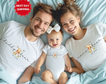 May include: A white t-shirt with the text "daddy bear est. 2024" and a cartoon image of Winnie the Pooh holding a jar of honey. A white t-shirt with the text "mama bear est. 2024" and a cartoon image of Winnie the Pooh holding a jar of honey. A white onesie with the text "baby bear est. 2024" and a cartoon image of Winnie the Pooh holding a jar of honey.