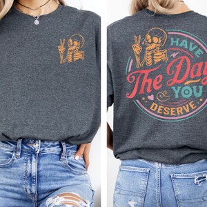 May include: A dark gray t-shirt with a skeleton graphic on the front and the text "Have the Day You Deserve" in a circle on the back. The text is in a colorful retro font.