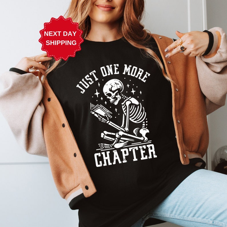 May include: Black t-shirt with a white graphic of a skeleton reading a book. The graphic says "Just One More Chapter".