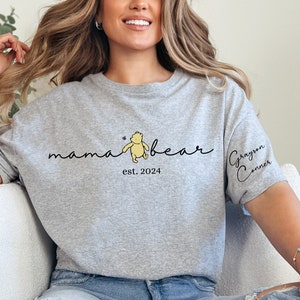 May include: Gray heather gray t-shirt with a Winnie the Pooh inspired graphic that says "mama bear est. 2024" and a small butterfly. The right sleeve has the name "Grayson Conner" written in cursive.