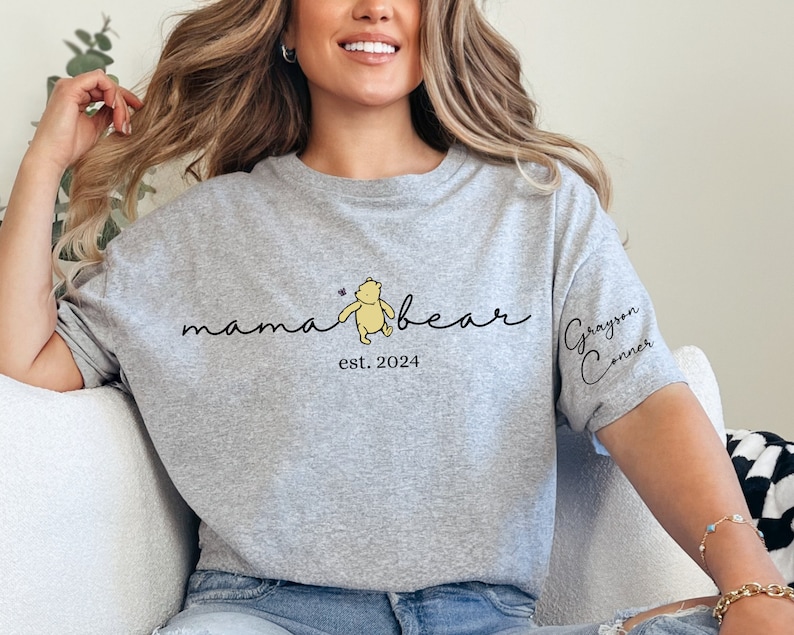 May include: Gray heather gray t-shirt with a Winnie the Pooh inspired graphic that says "mama bear est. 2024" and a small butterfly. The right sleeve has the name "Grayson Conner" written in cursive.