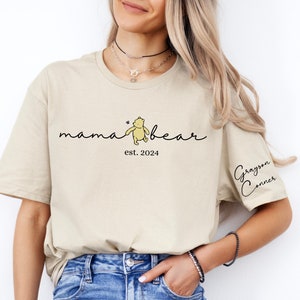 May include: A beige t-shirt with the text "mama bear" and a small illustration of Winnie the Pooh. The text "est. 2024" is below the bear. The text "Grayson Conner" is on the right sleeve.