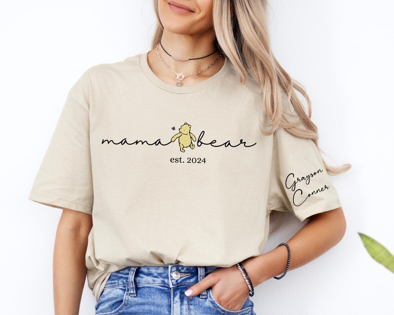 May include: A beige t-shirt with the text "mama bear" and a small illustration of Winnie the Pooh. The text "est. 2024" is below the bear. The text "Grayson Conner" is on the right sleeve.