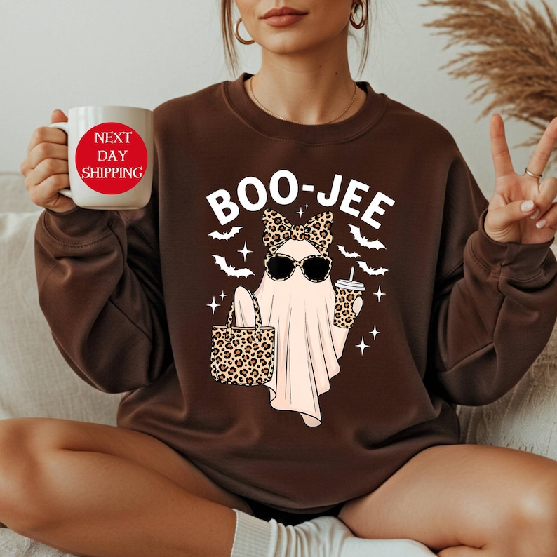 May include: Brown sweatshirt with a Halloween-themed graphic. The graphic features a ghost wearing sunglasses, a bow, and holding a leopard print bag and coffee cup. The text "BOO-JEE" is above the ghost. A white mug with red text is held in the hand.