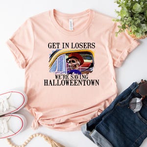 May include: A pink t-shirt with the text "Get in losers, we're saving Halloweentown" and a cartoon skeleton driving a car.