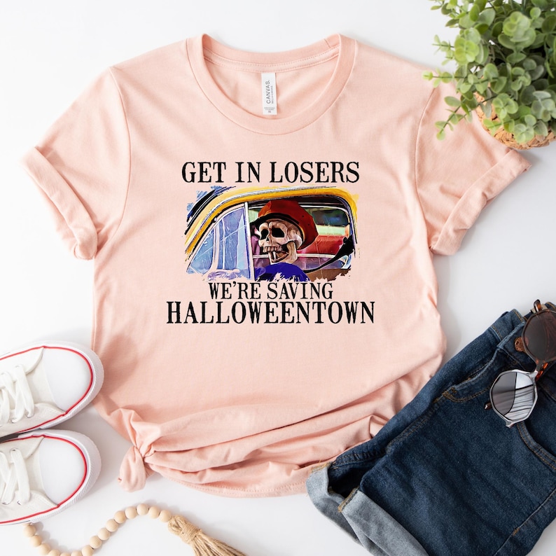 May include: A pink t-shirt with the text "Get in losers, we're saving Halloweentown" and a cartoon skeleton driving a car.