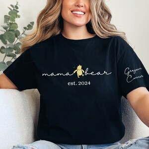 May include: Black t-shirt with a white graphic that says "mama bear" with a small Winnie the Pooh illustration and "est. 2024". The right sleeve has the name "Grayson Conner" written in cursive.