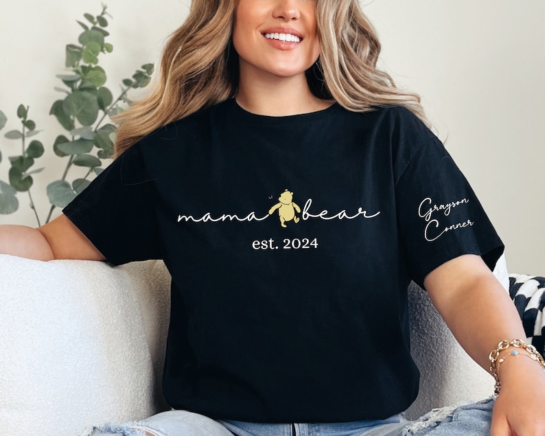 May include: Black t-shirt with a white graphic that says "mama bear" with a small Winnie the Pooh illustration and "est. 2024". The right sleeve has the name "Grayson Conner" written in cursive.