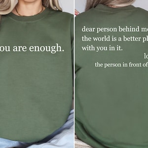 Puede incluir: Una sudadera verde con el texto "you are enough." en la parte delantera y el texto "dear person behind me, the world is a better place with you in it. love, the person in front of you" en la parte trasera.