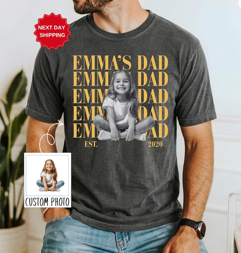 Puede incluir: Camiseta gris oscuro con una foto personalizada y el texto "EMMA'S DAD" en dorado. La camiseta presenta una imagen en blanco y negro de un ni&ntilde;o y el texto "EST. 2020".