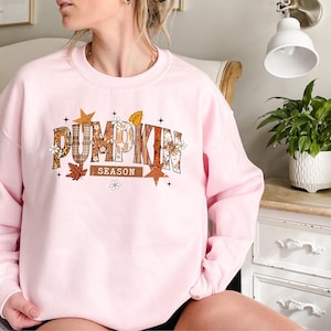 Może przedstawiać: Jasnor&oacute;żowa bluza z napisem "PUMPKIN SEASON" w jesiennym wzornictwie. Tekst zawiera wz&oacute;r w kratę, kwiatowe akcenty i jesienne liście. Bluza ma okrągły dekolt.