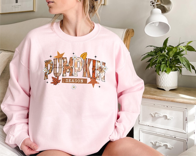 Może przedstawiać: Jasnor&oacute;żowa bluza z napisem "PUMPKIN SEASON" w jesiennym wzornictwie. Tekst zawiera wz&oacute;r w kratę, kwiatowe akcenty i jesienne liście. Bluza ma okrągły dekolt.