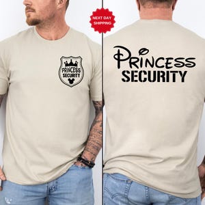 Może przedstawiać: Beżowa koszulka z napisem "Princess Security" w kolorze czarnym z przodu i z tyłu. Przedni wzór to tarcza z koroną i uszami Myszki Miki. Tył ma ten sam tekst w większej czcionce.