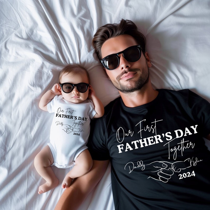 May include: A black t-shirt with the text "Our First Father's Day Together" and a hand-holding graphic. The text "Daddy" and "Kylan" are written above the hands. The year "2024" is written below the hands. A white onesie with the text "Our First Father's Day Together" and a hand-holding graphic. The text "Dad" and "Son" are written above the hands. The year "2024" is written below the hands.