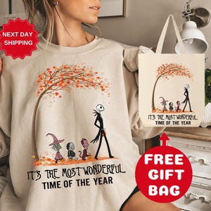 Op de afbeelding: Een beige sweatshirt met een afbeelding van Jack Skellington en andere personages uit The Nightmare Before Christmas. De afbeelding bevat een herfstboom met oranje en rode bladeren. De tekst "It's the most wonderful time of the year" staat op het sweatshirt gedrukt. Een bijpassende tote bag is ook inbegrepen.