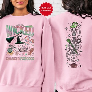 Puede incluir: Sudadera rosa claro con la palabra "WICKED" en verde y azul, junto con ilustraciones tem&aacute;ticas de brujas. La parte trasera de la sudadera presenta un dise&ntilde;o vertical con un sombrero de bruja, estrellas y s&iacute;mbolos m&aacute;gicos.