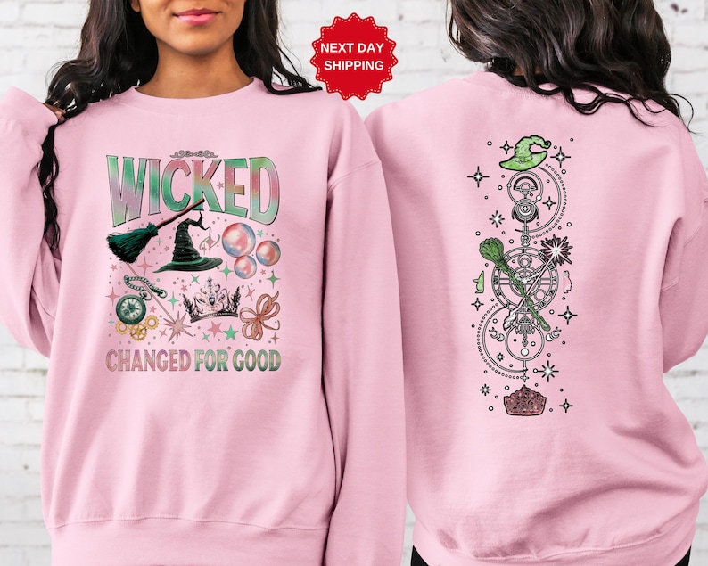 Puede incluir: Sudadera rosa claro con la palabra "WICKED" en verde y azul, junto con ilustraciones tem&aacute;ticas de brujas. La parte trasera de la sudadera presenta un dise&ntilde;o vertical con un sombrero de bruja, estrellas y s&iacute;mbolos m&aacute;gicos.