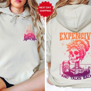 May include: A light beige hoodie with a graphic of a skeleton holding a coffee cup and the text "Expensive and Talks Back" on the back. The front of the hoodie has a graphic of a skeleton hand with roses.