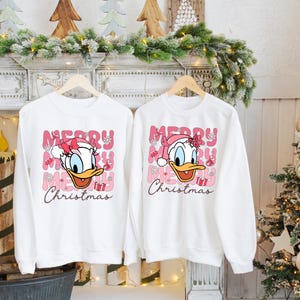 May include: Two white sweatshirts with a pink and white Christmas design featuring a cartoon duck wearing a Santa hat. The text on the sweatshirts reads "Merry Christmas".