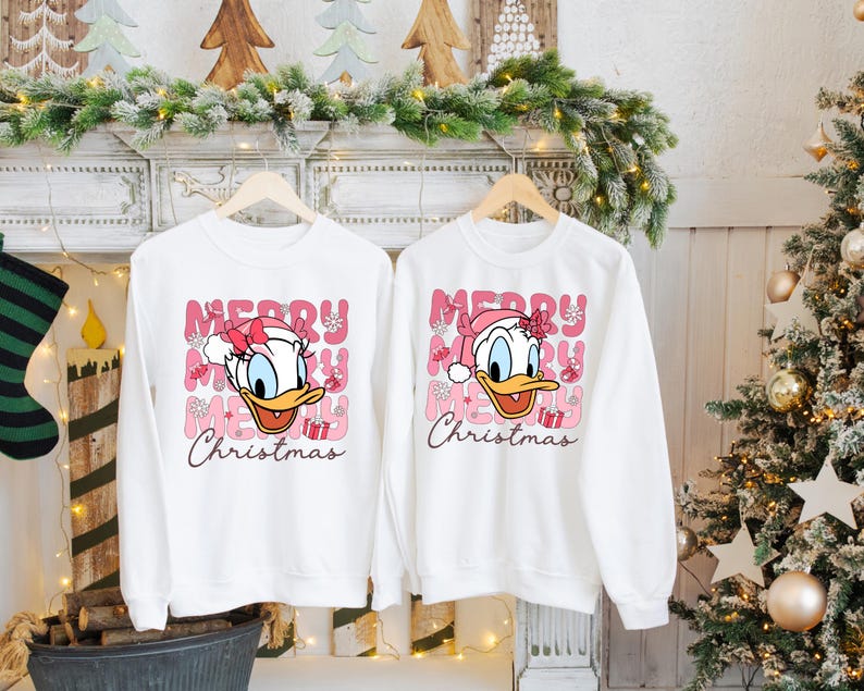 May include: Two white sweatshirts with a pink and white Christmas design featuring a cartoon duck wearing a Santa hat. The text on the sweatshirts reads "Merry Christmas".